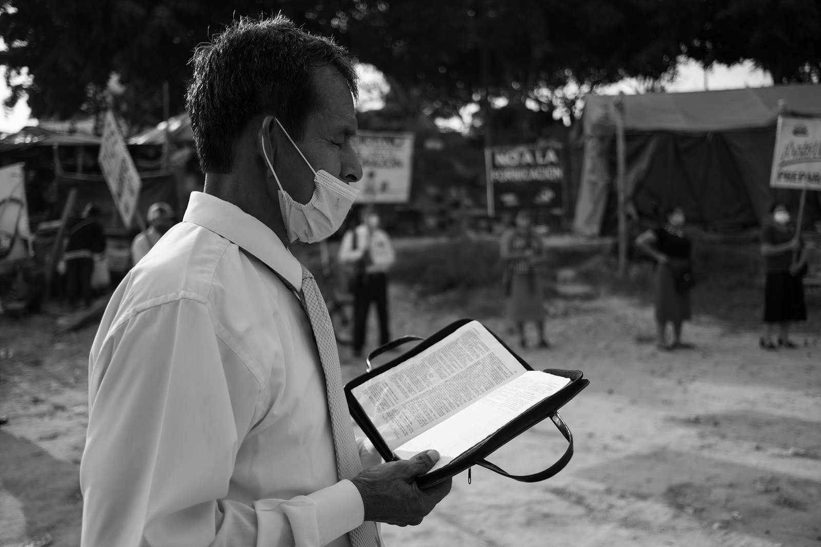 A preacher is standing holding a Bible, with his eyes closed and a medical mask on.