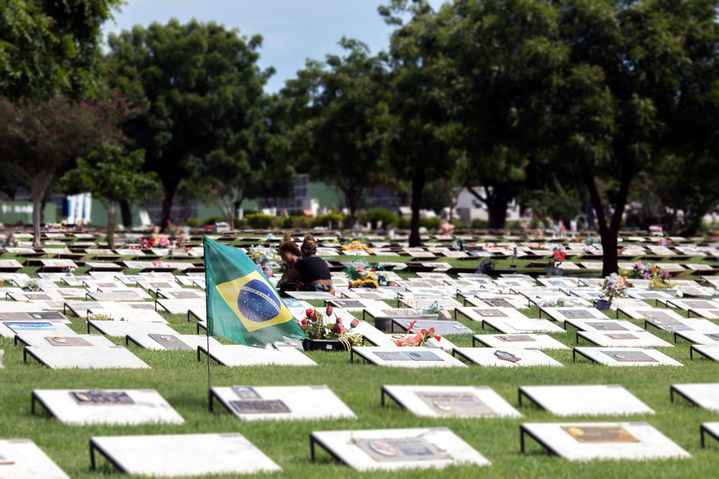 Um campo de cemitério com fileiras de marcadores de sepulturas. Algumas das sepulturas têm flores. Uma bandeira brasileira ondula em primeiro plano. Duas pessoas visitam uma sepultura em segundo plano.